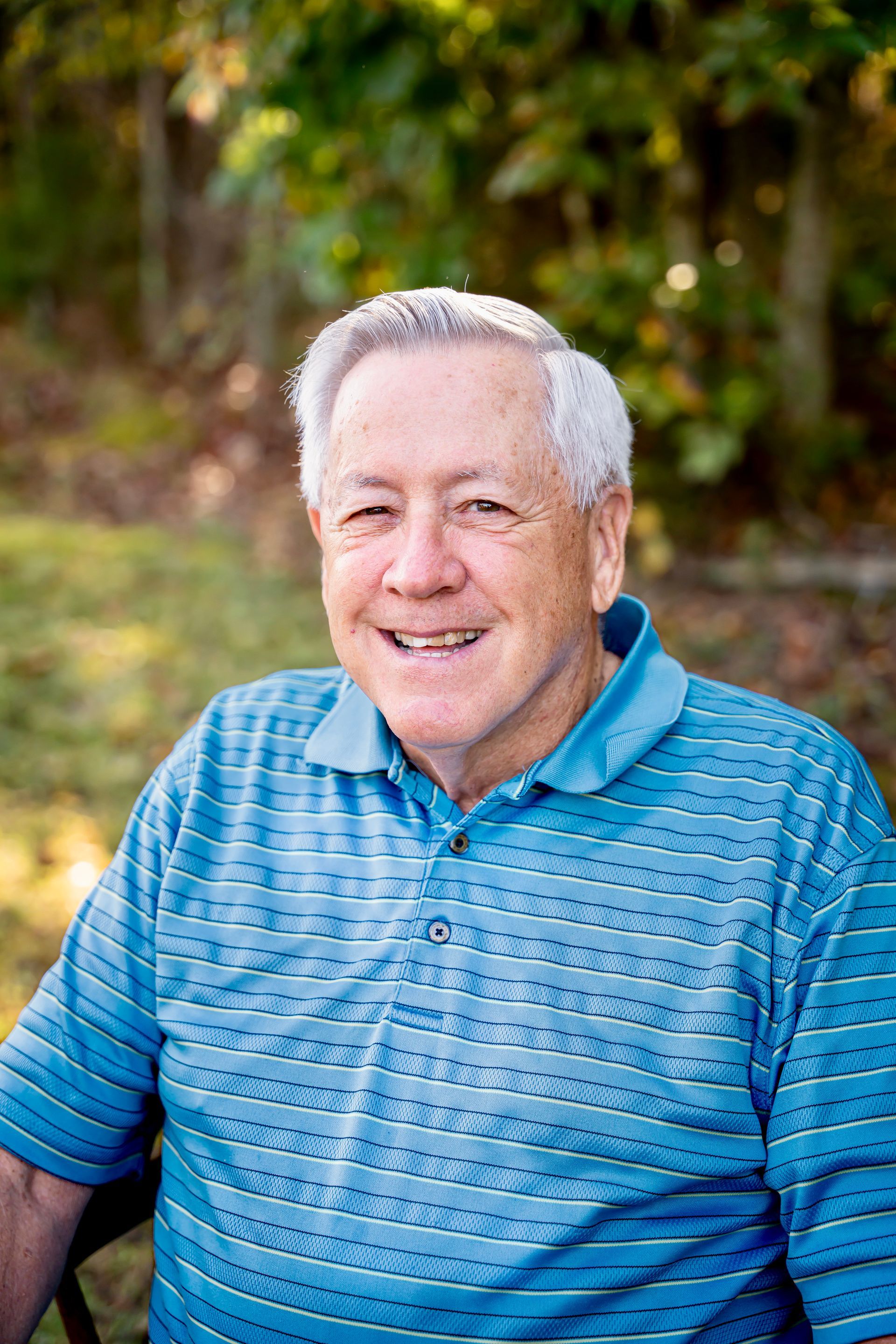 Man with white hair smiles, wearing a blue polo shirt, outdoors with blurred green foliage.