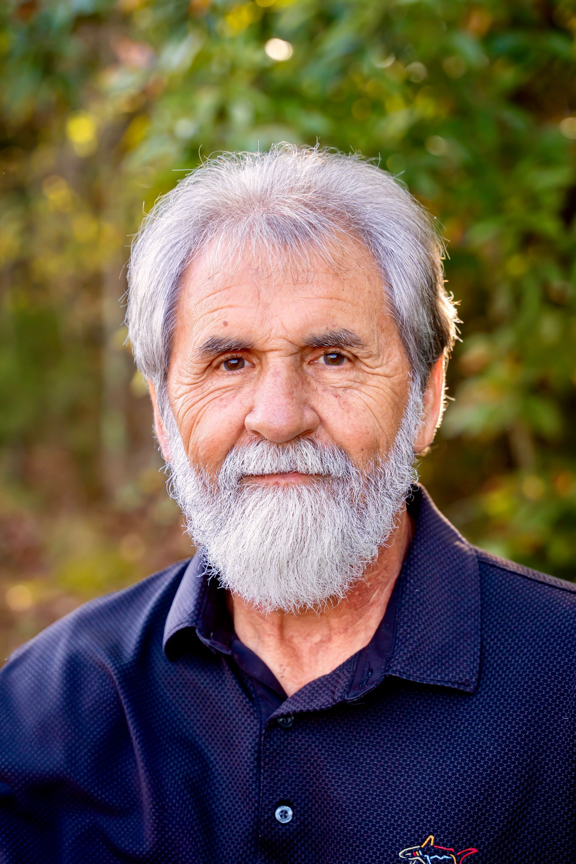 Man with gray beard, wearing a navy shirt, smiles outdoors with a green and yellow blurred background.