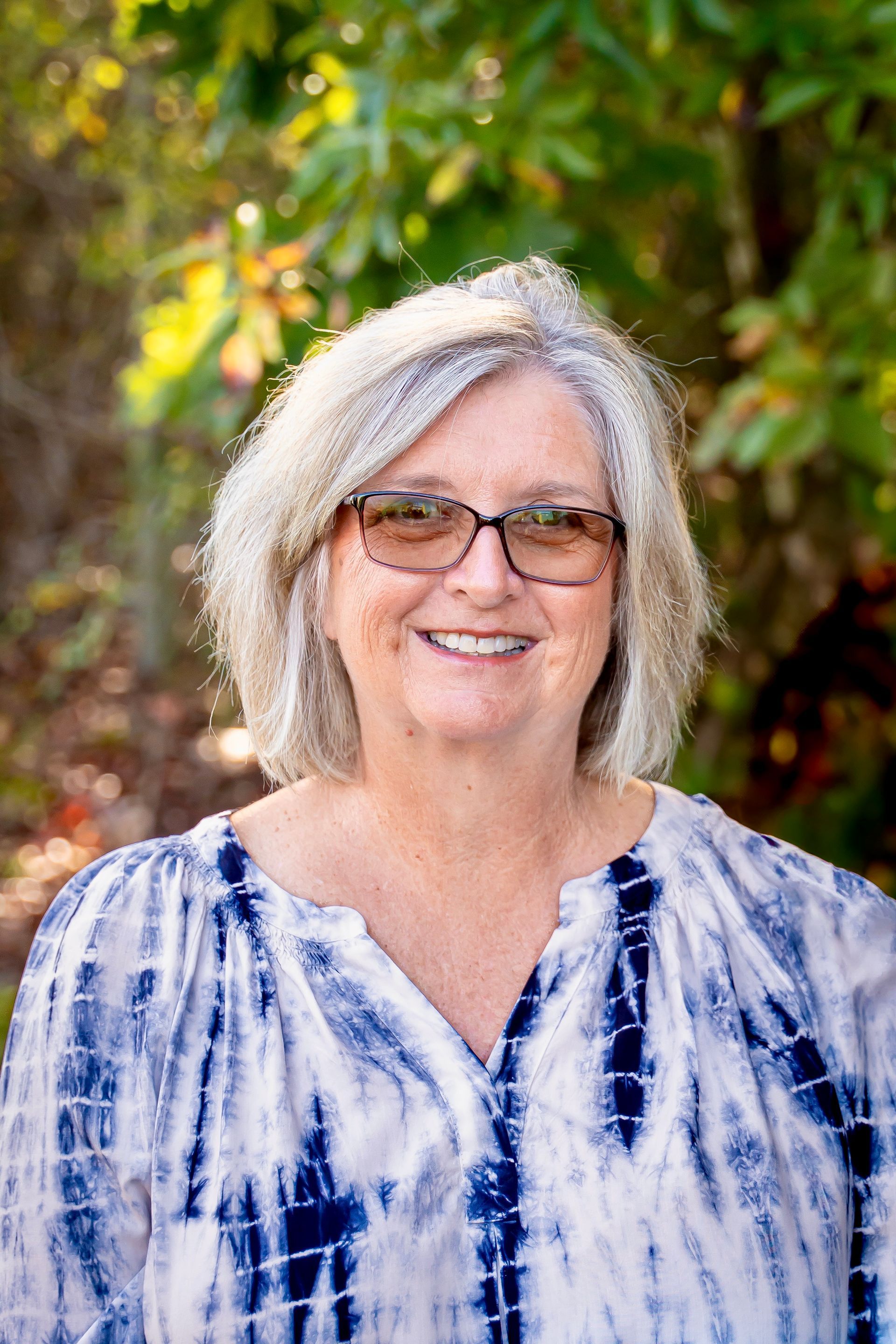 Woman with glasses smiles; outdoors, wearing tie-dye top with neutral background.