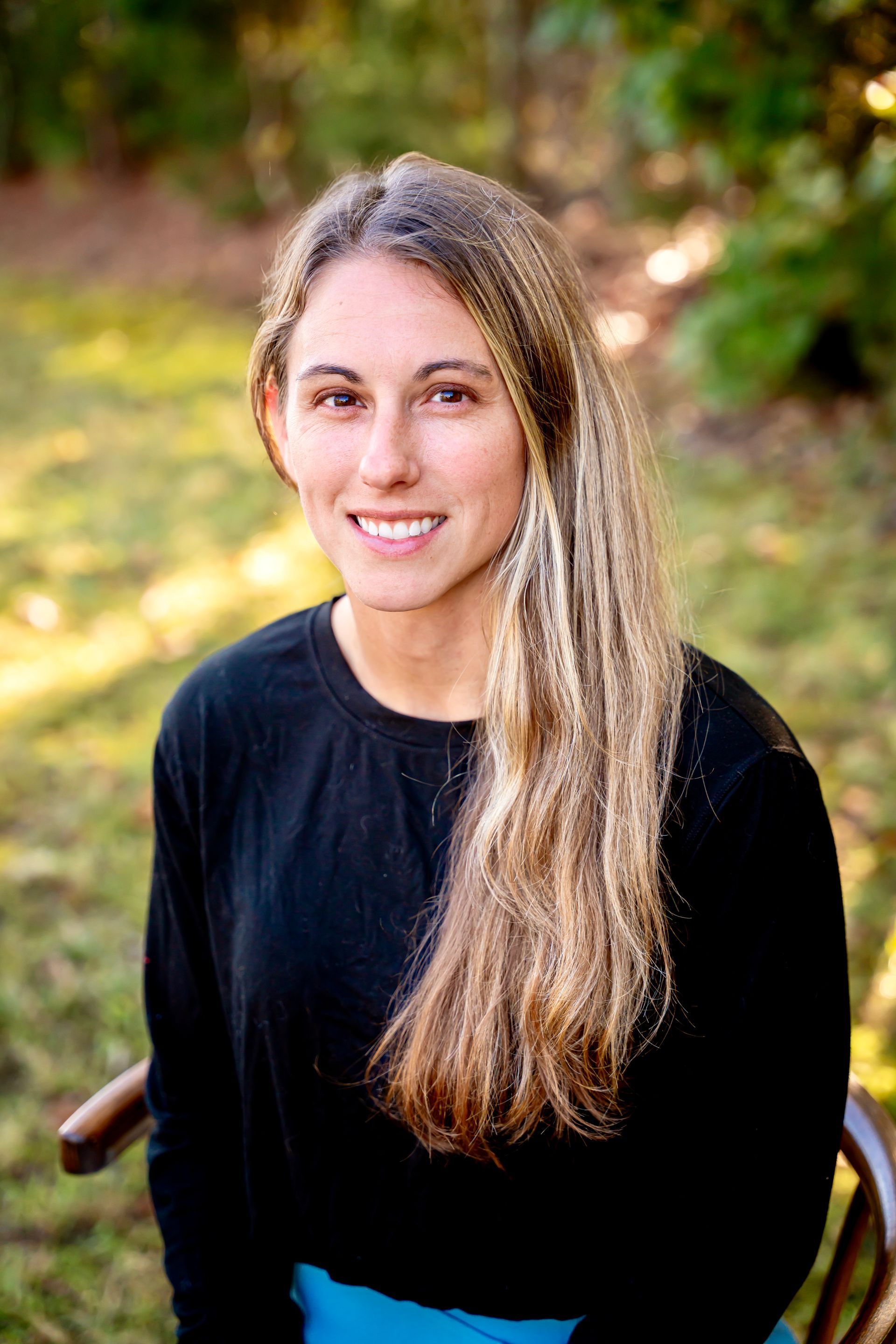 Woman with long blonde hair smiles outdoors, wearing black shirt and blue pants.