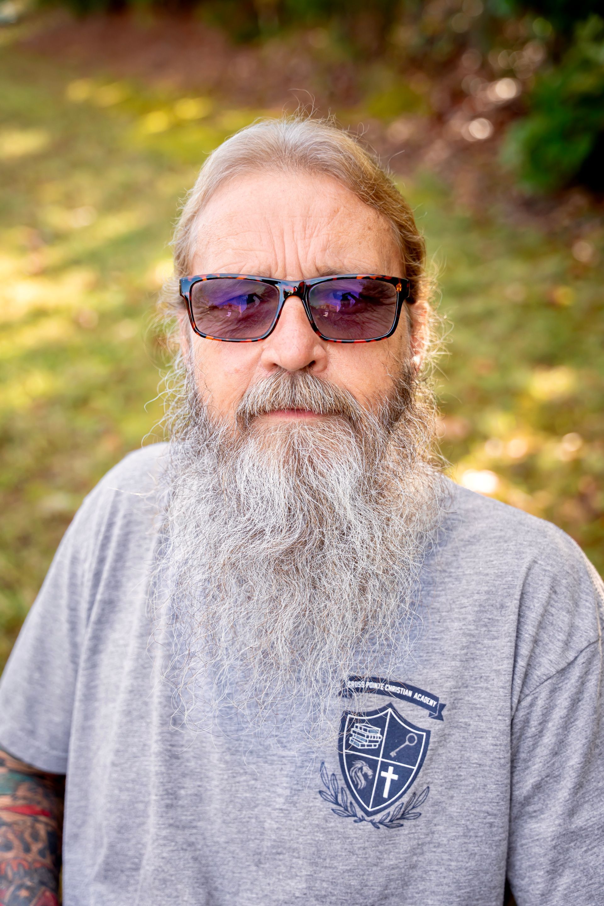 Man with long gray beard and sunglasses outdoors. Gray t-shirt with a crest.