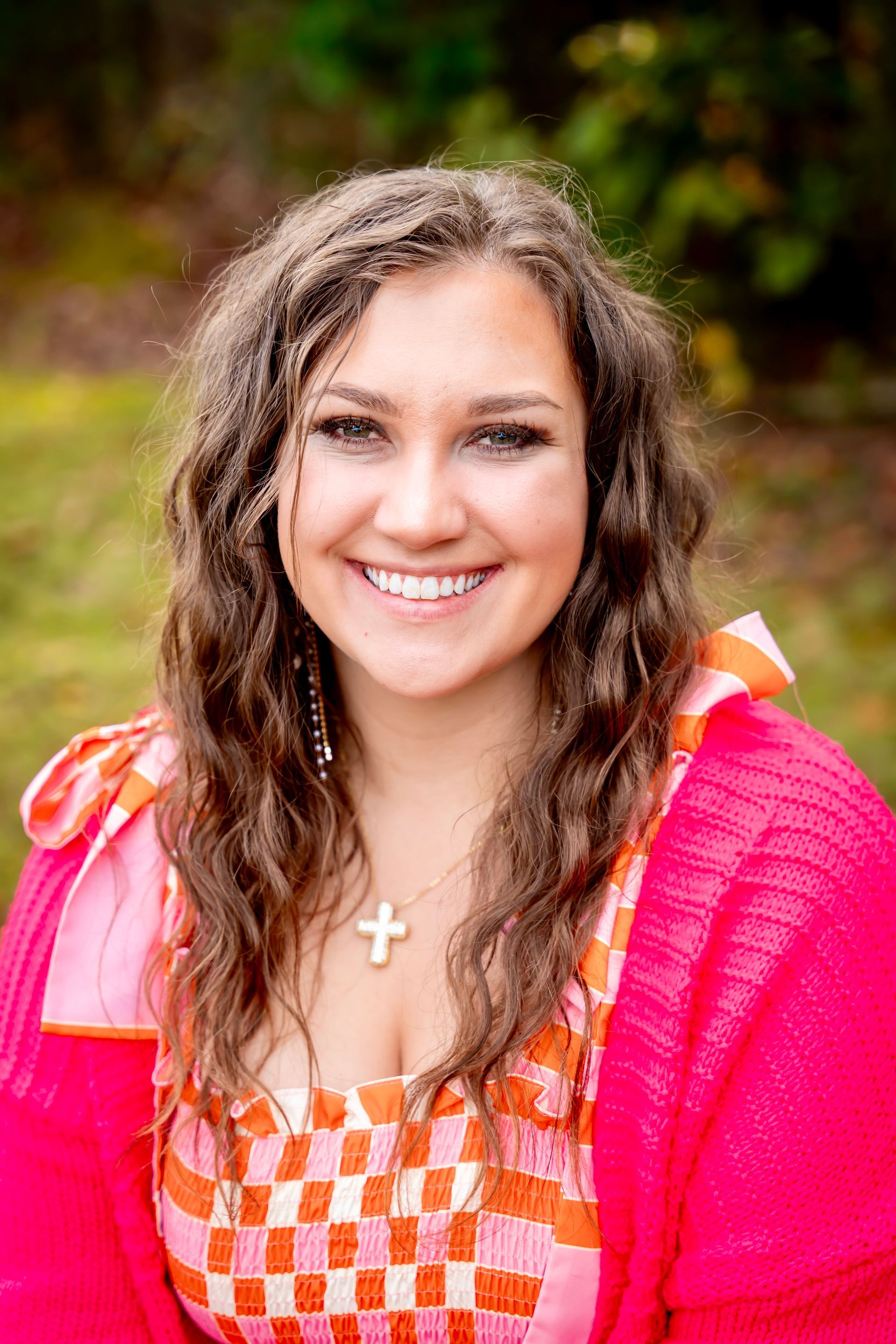 Woman smiling, wearing pink sweater over patterned top, gold cross necklace.