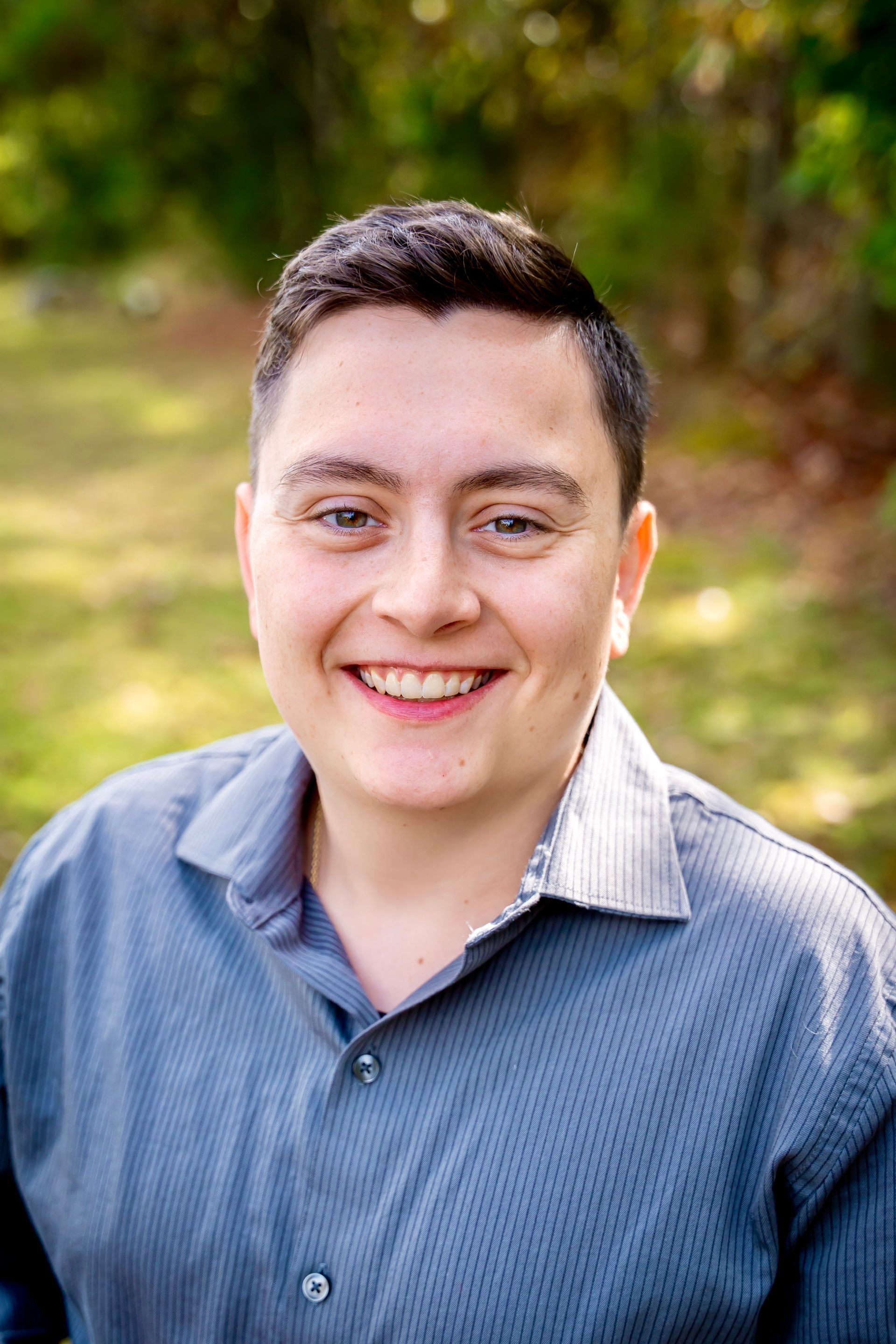 Person smiling, wearing a blue button-up shirt, against a blurred green background.