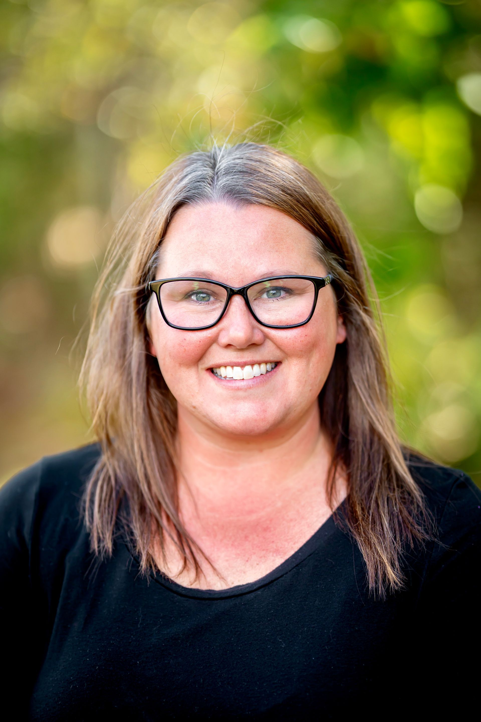 Woman with glasses smiles; outdoors, blurred green backdrop, wearing black top.