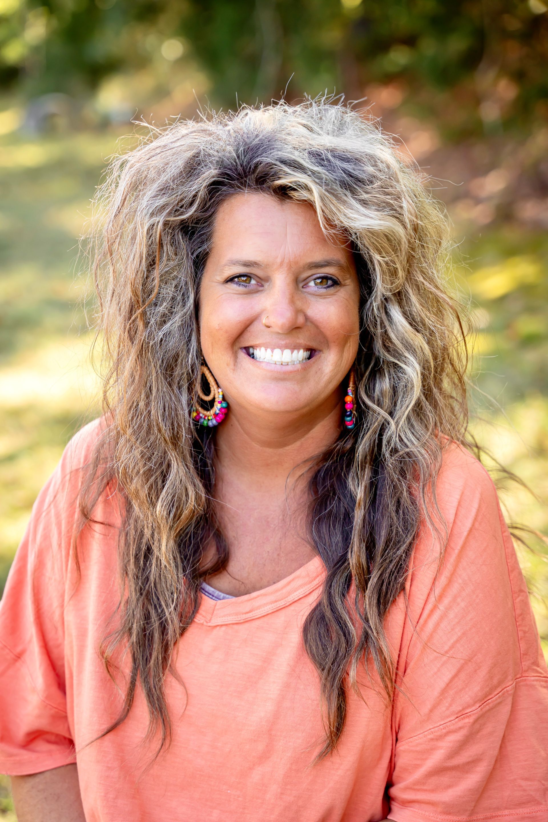 Woman with long curly hair smiles outdoors, wearing an orange top and hoop earrings.