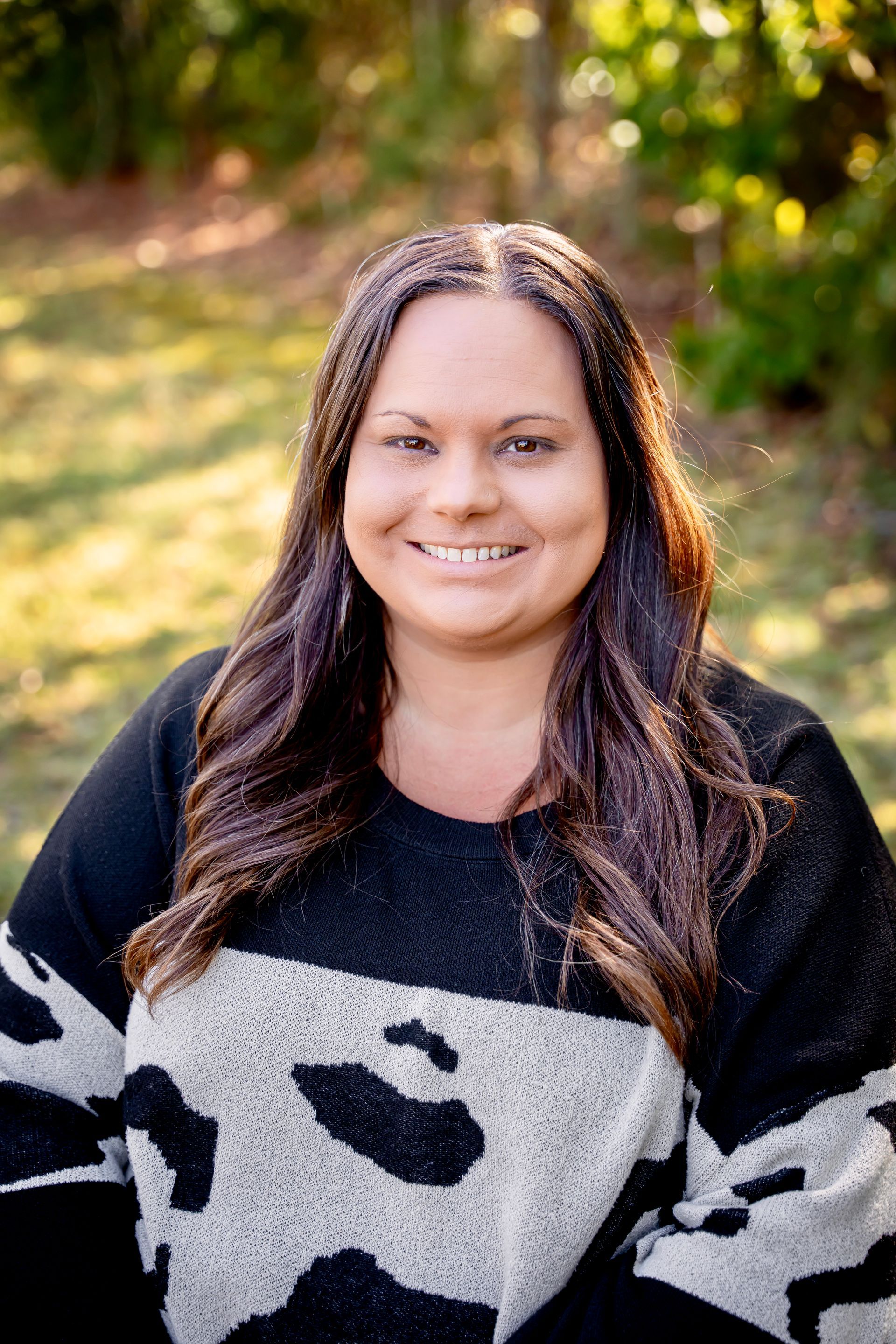 Woman with long brown hair smiles, wearing black and white patterned sweater outdoors.