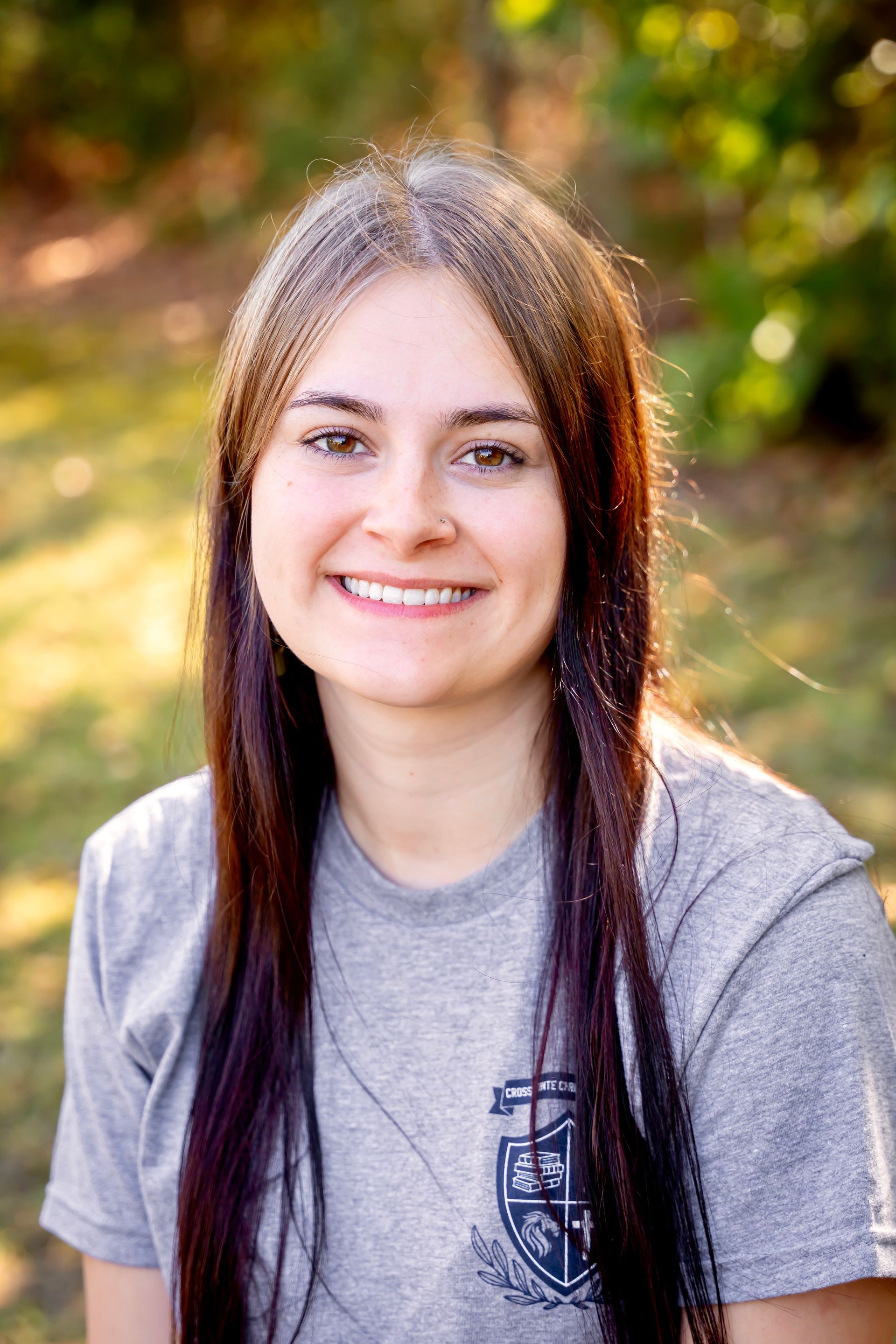 Woman with long brown hair smiles outdoors in a grey shirt.