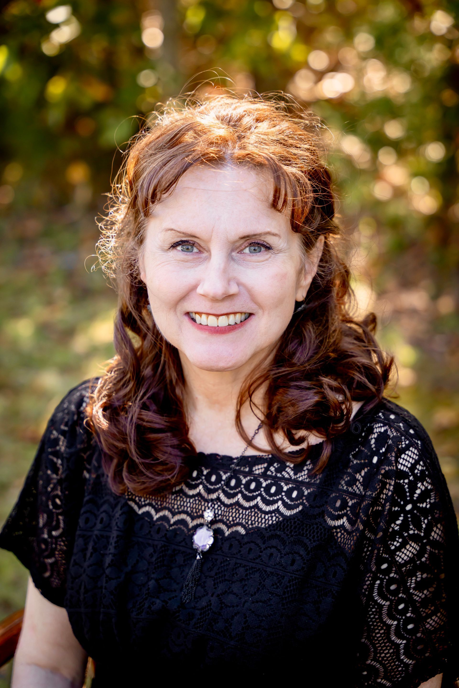 Woman with reddish-brown curly hair smiles at camera, wearing a black lace top outside with blurred foliage.