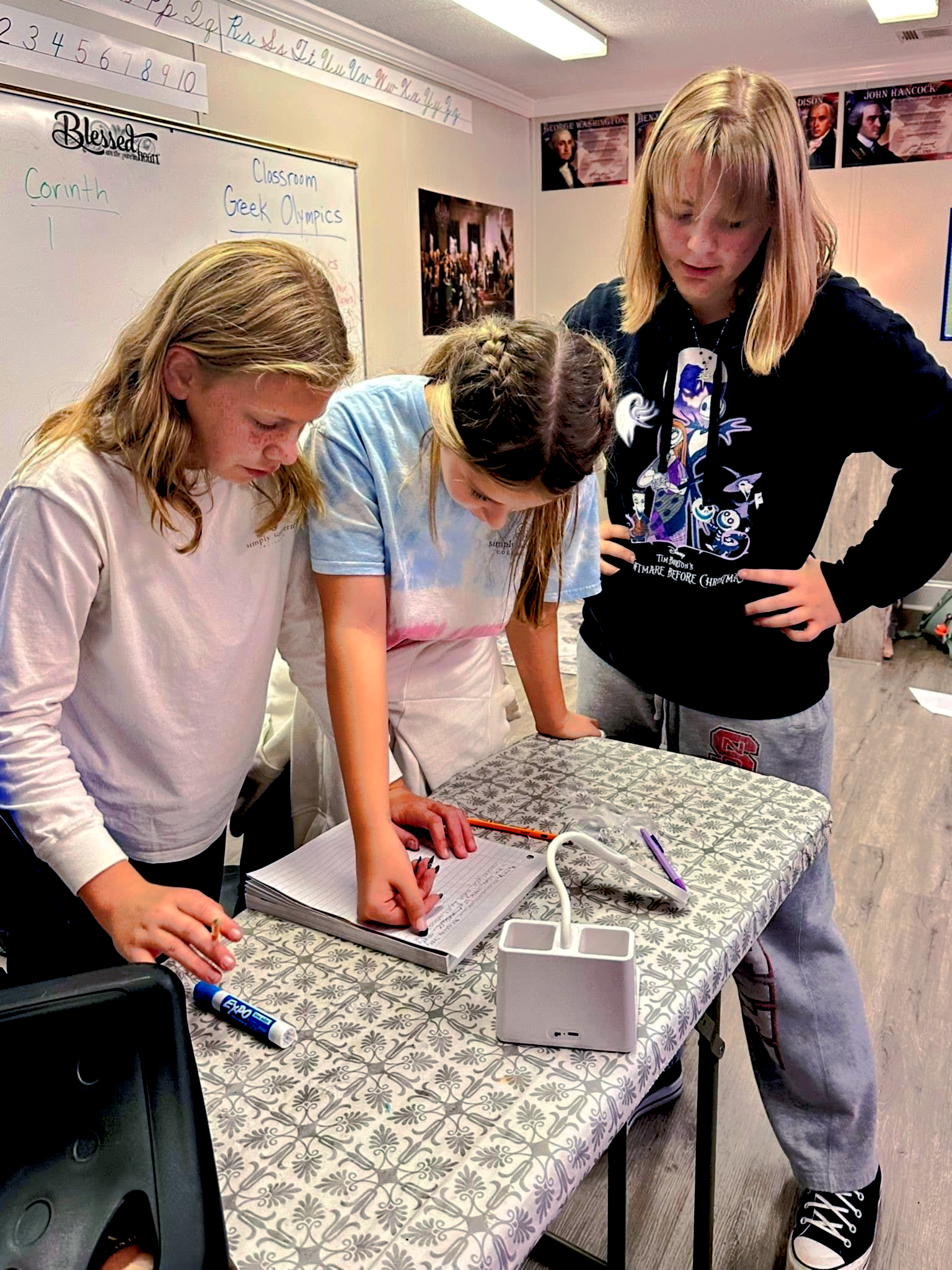Three students in a classroom examine a notebook on a table. One writes with a pencil.