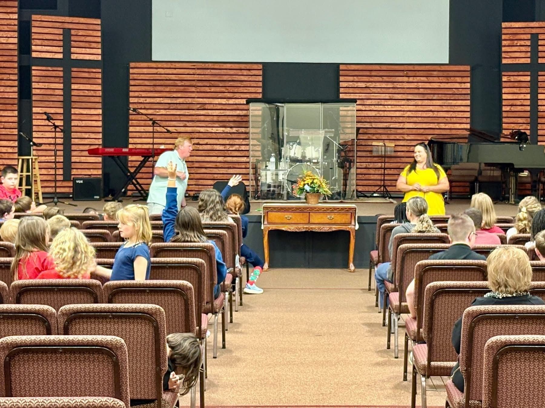 People in a church auditorium, two speakers on stage with a wooden backdrop.