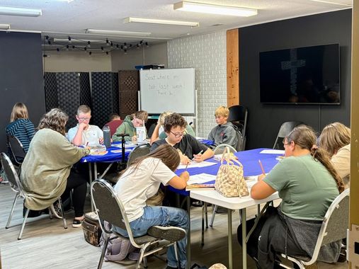 People seated around tables writing in a classroom. A whiteboard and TV are visible.