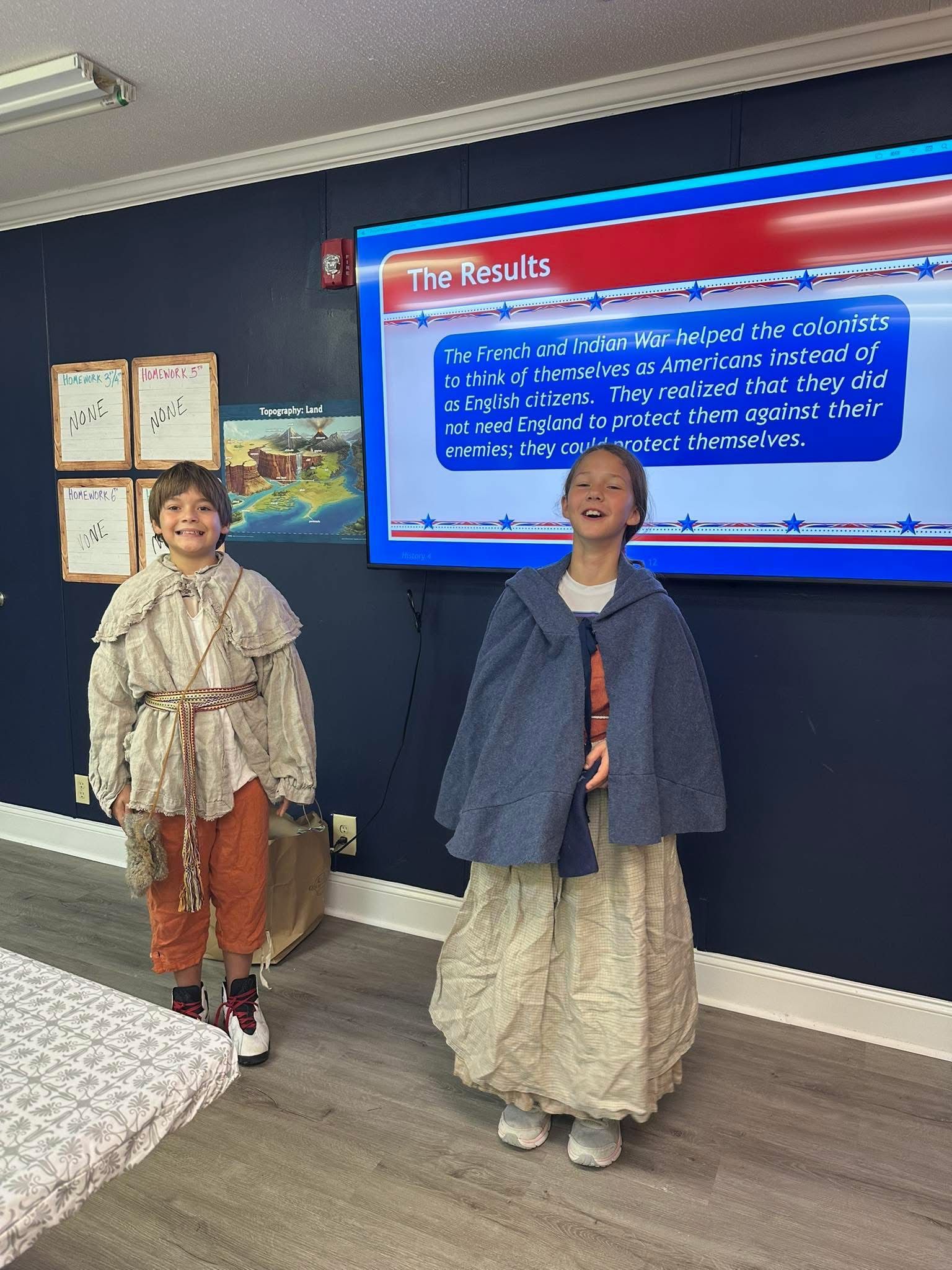 Two children in period costumes stand near a screen displaying text about the Puritans.