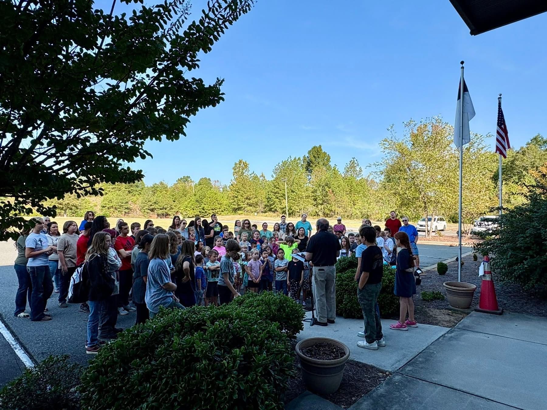 Group of people gathered outside, some listening to a speaker, with flags and trees in the background.