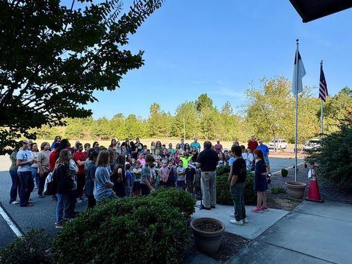 Group of people gathered outside, some listening to a speaker, with flags and trees in the background.