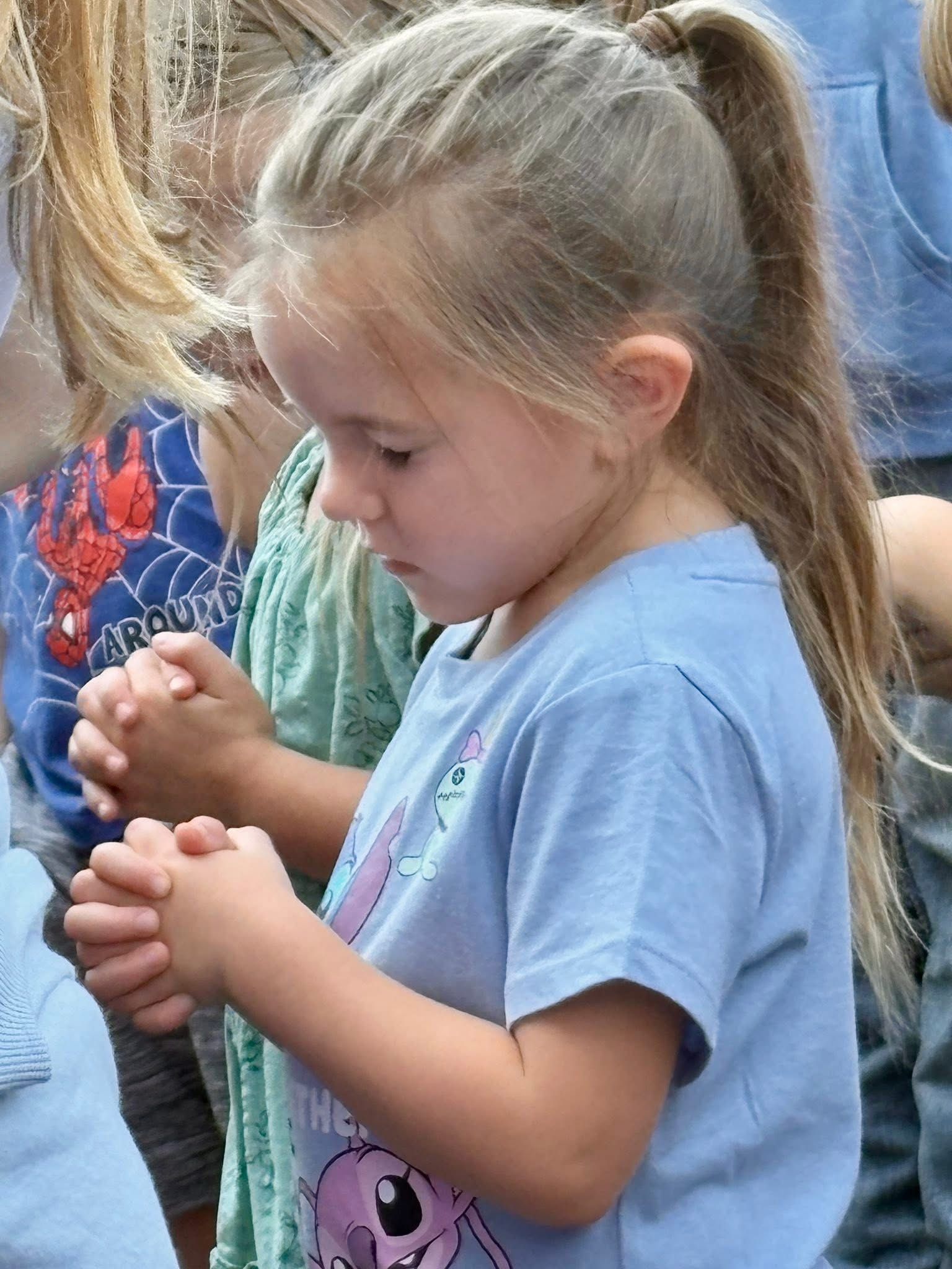 Girl with blonde hair in a ponytail, clasped hands, eyes closed, wearing a blue shirt.