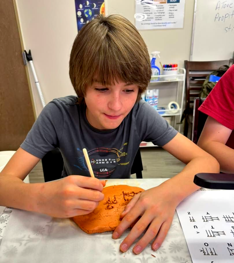 Boy using a tool to create marks on a clay-like orange disc. He sits at a table with papers, focused expression.