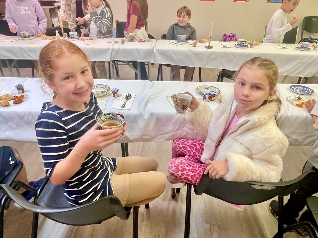 Two girls smiling at a table, one holding a drink. Other children at a party table with food and decorations.