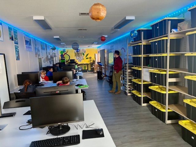 A computer lab with students and a teacher. Shelves hold storage bins. Blue and yellow decor.