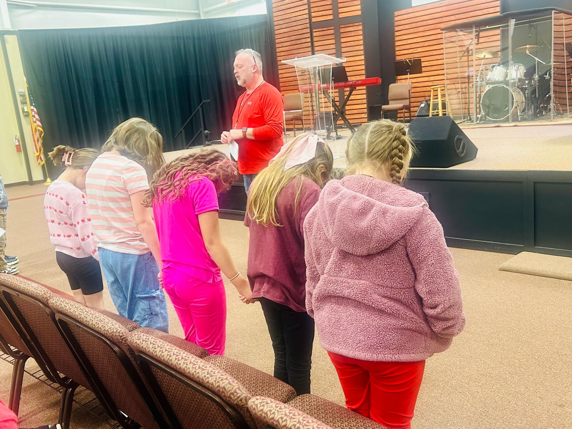 Children and adult in a church. A man in red leads children in prayer.