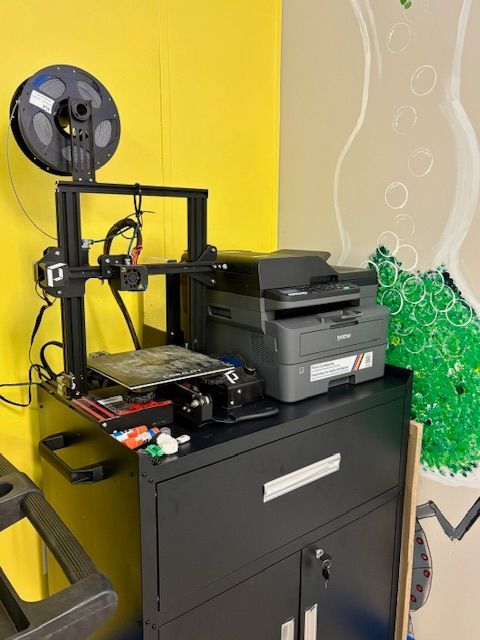 A 3D printer with a spool of filament next to a printer on a black cabinet against a yellow wall.