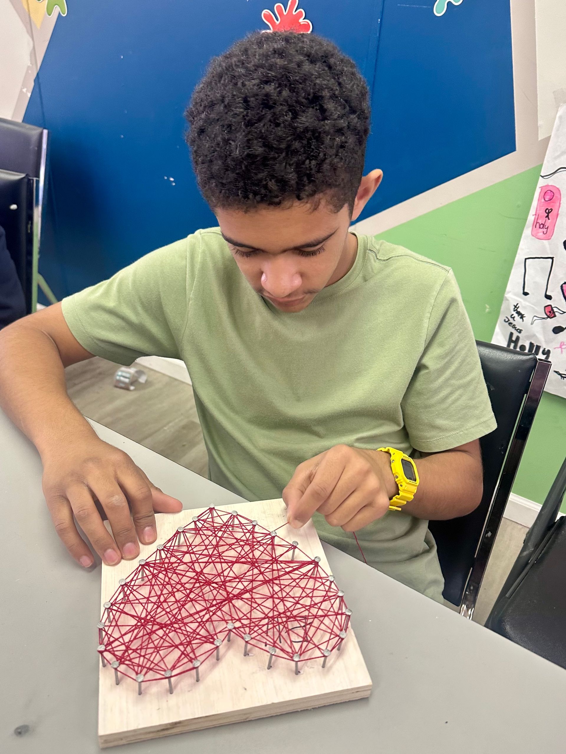Boy creating string art heart with red thread on a wooden board.