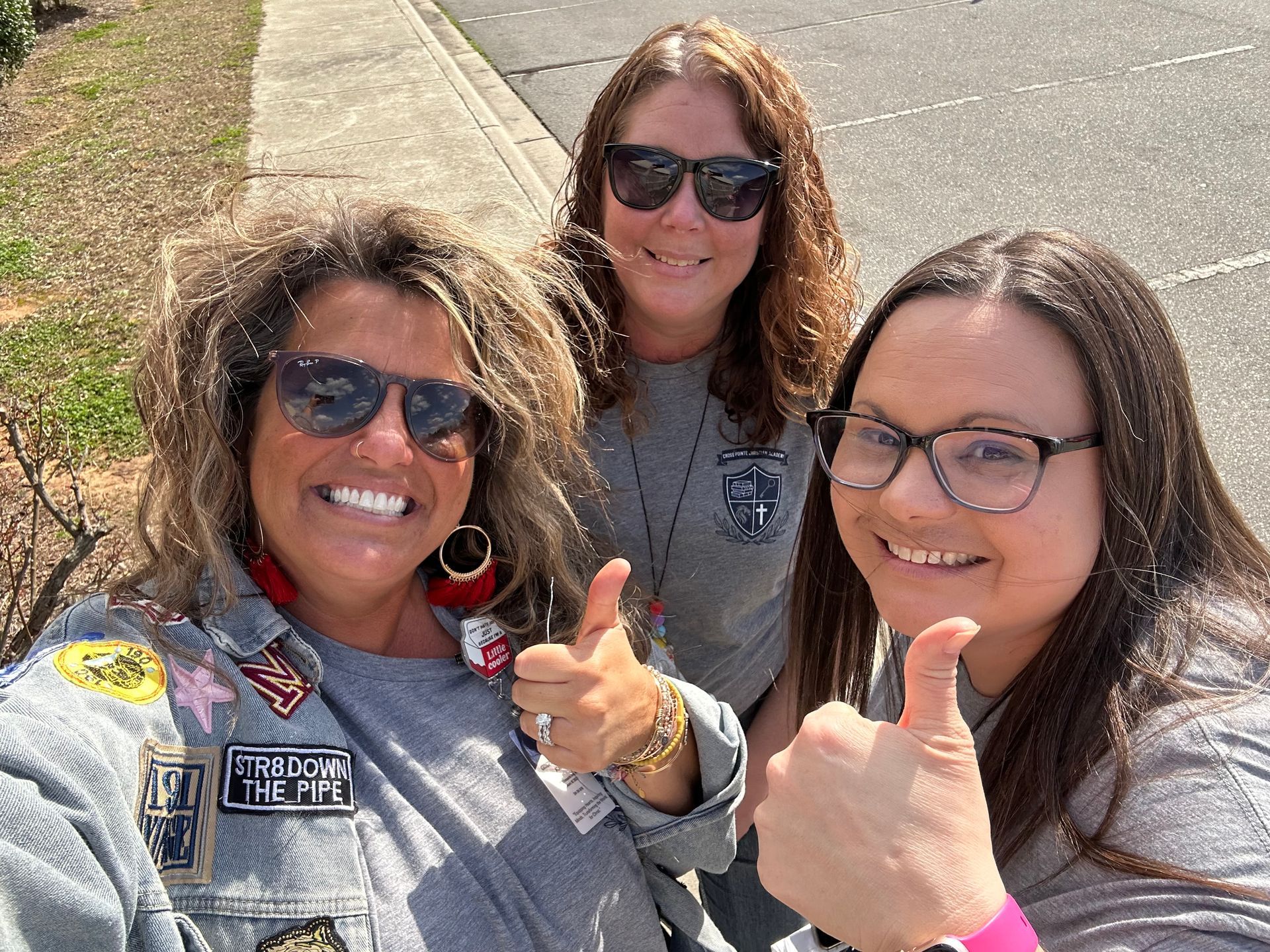 Three people smiling and giving thumbs up outdoors. They wear sunglasses and casual clothing.