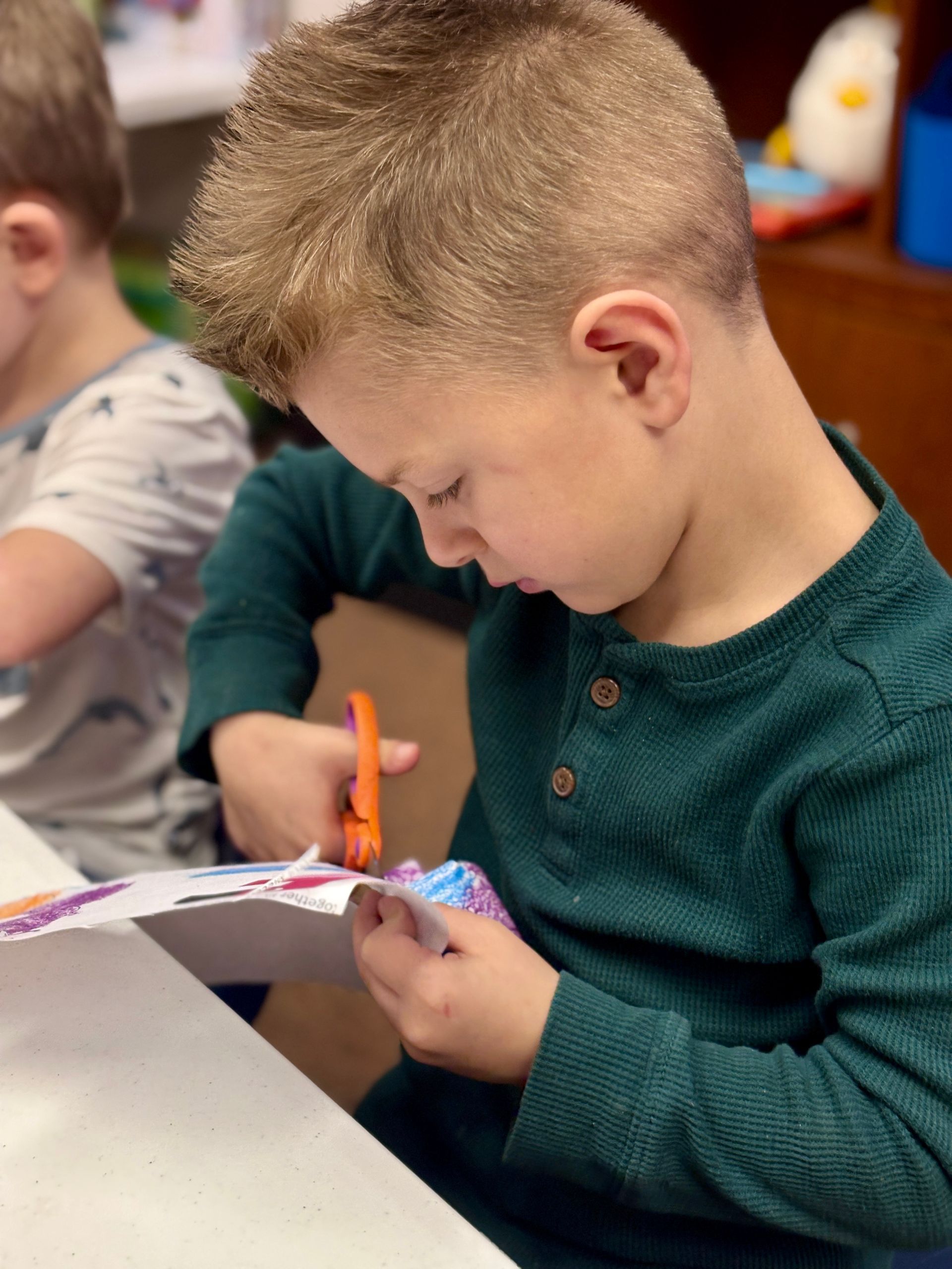 Boy in green shirt carefully cuts paper with orange scissors.