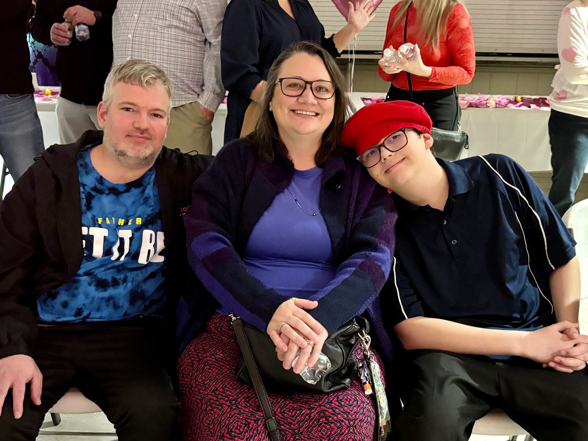 Three people seated together smiling: a woman in glasses, a man in a tie-dye shirt, and a person in a red cap.