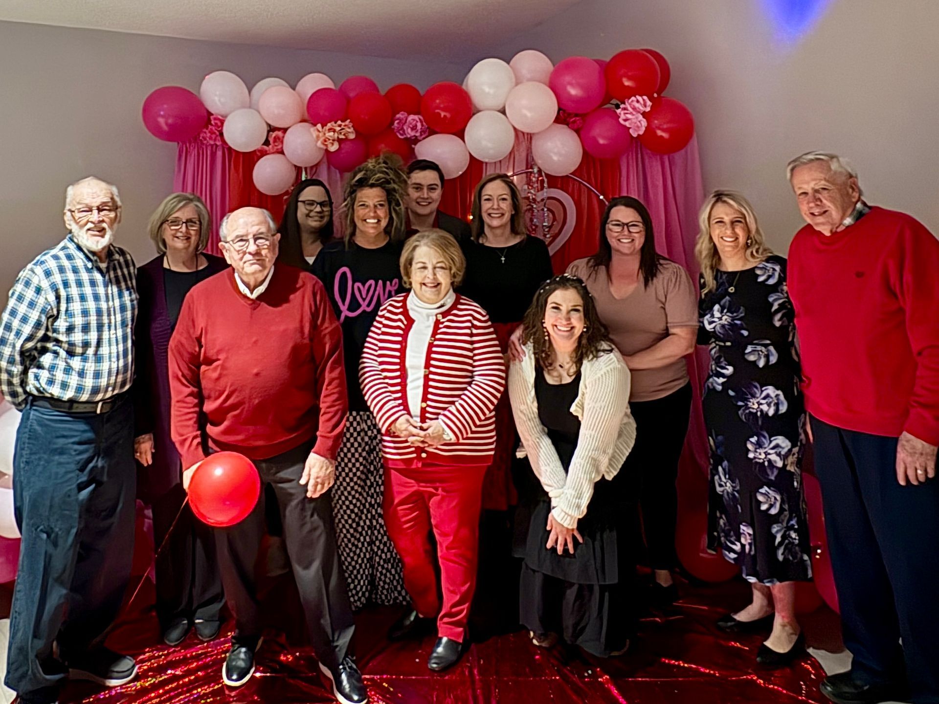 Group of people smiling in front of a Valentine's Day balloon backdrop. Red and pink colors dominate the scene.