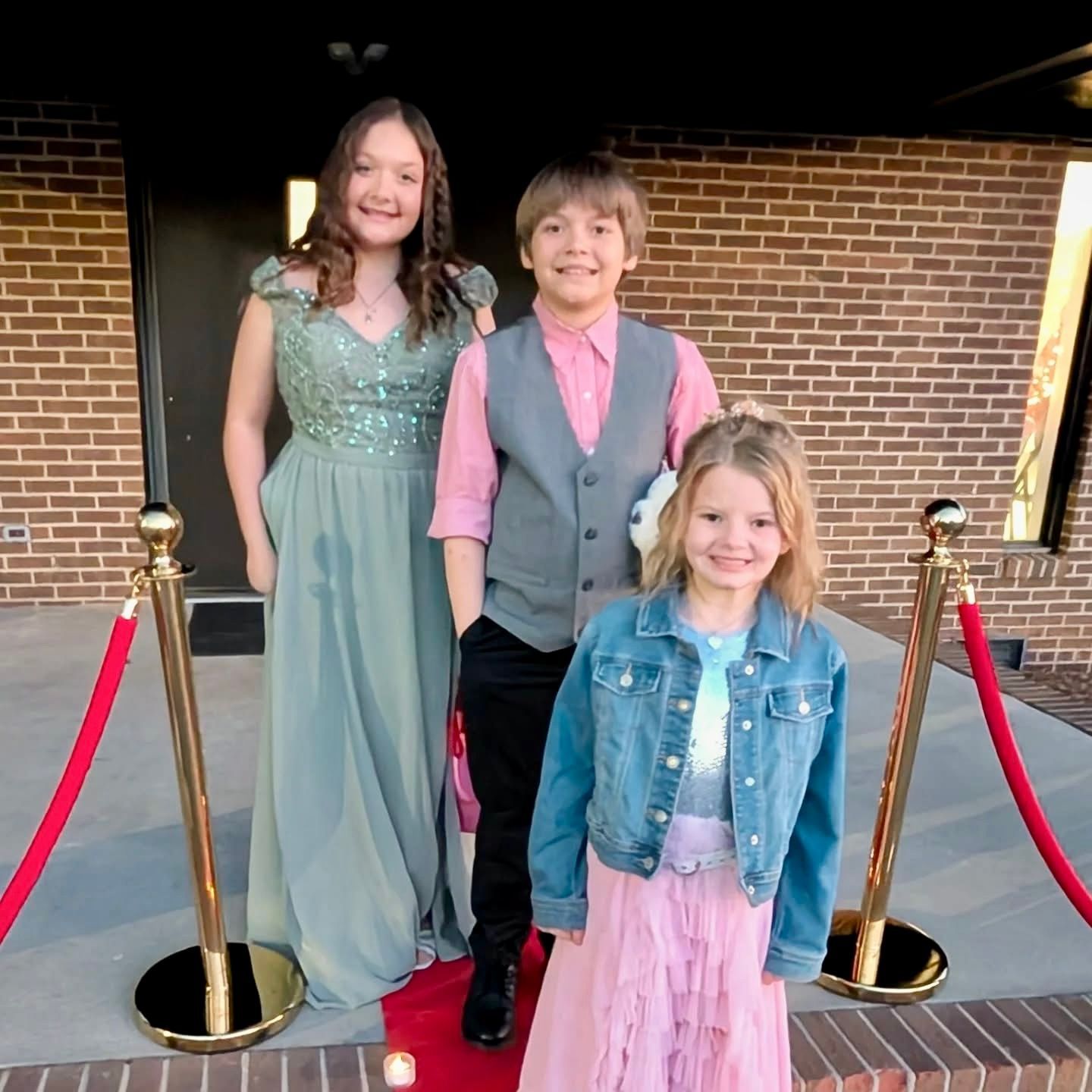 Three children pose on a red carpet. One girl in mint green, the boy in a pink shirt, the younger girl has a denim jacket.