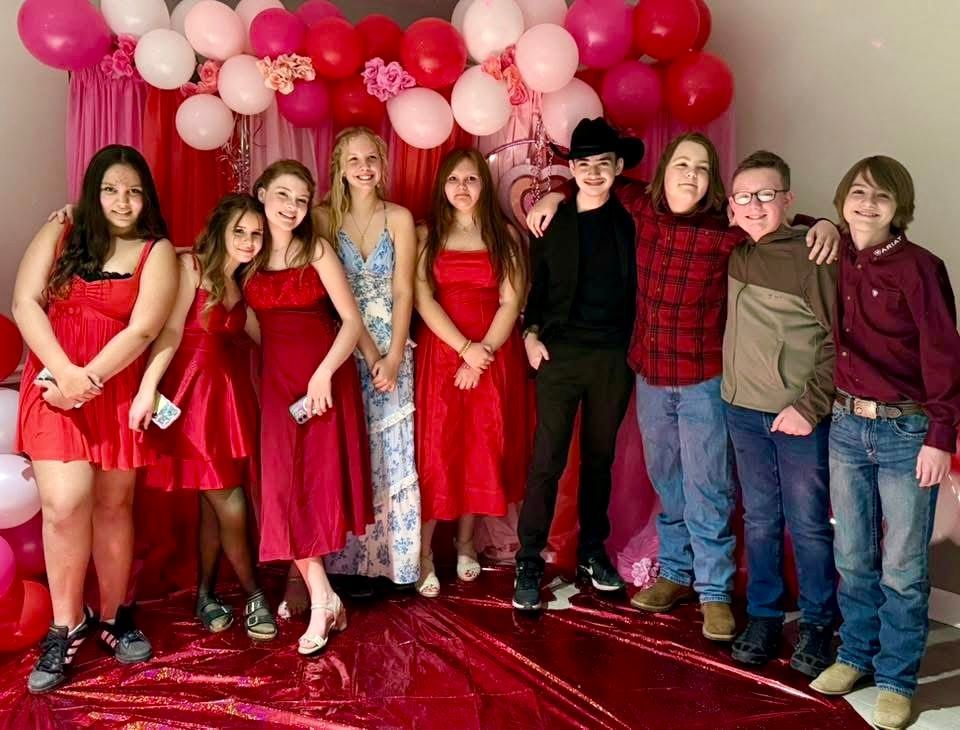 Group of teens posing in front of a Valentine's Day balloon backdrop; wearing red and pink clothing.