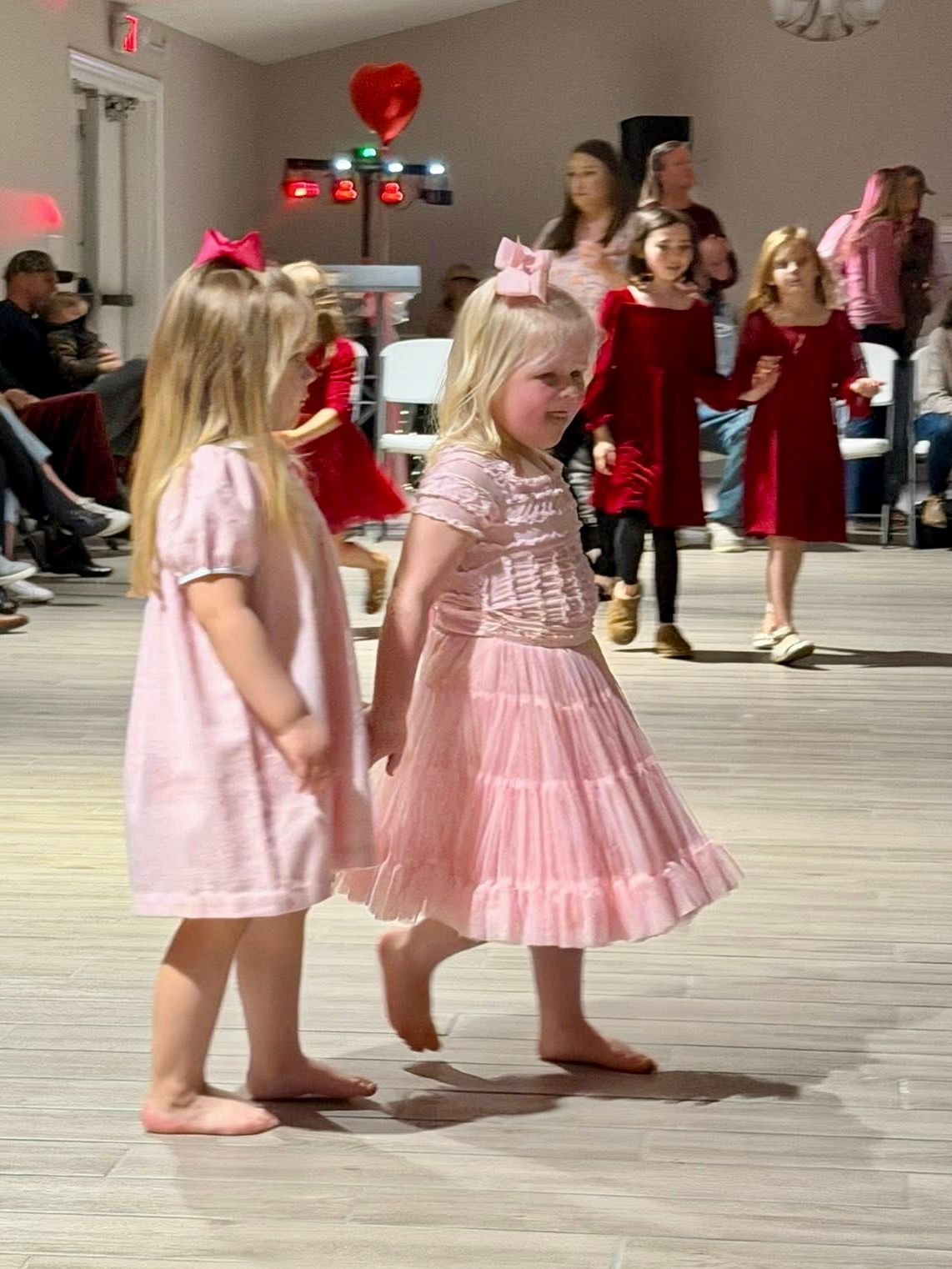 Two barefoot girls in pink dresses, one with a bow, walk on a dance floor. Other girls and people in the background.