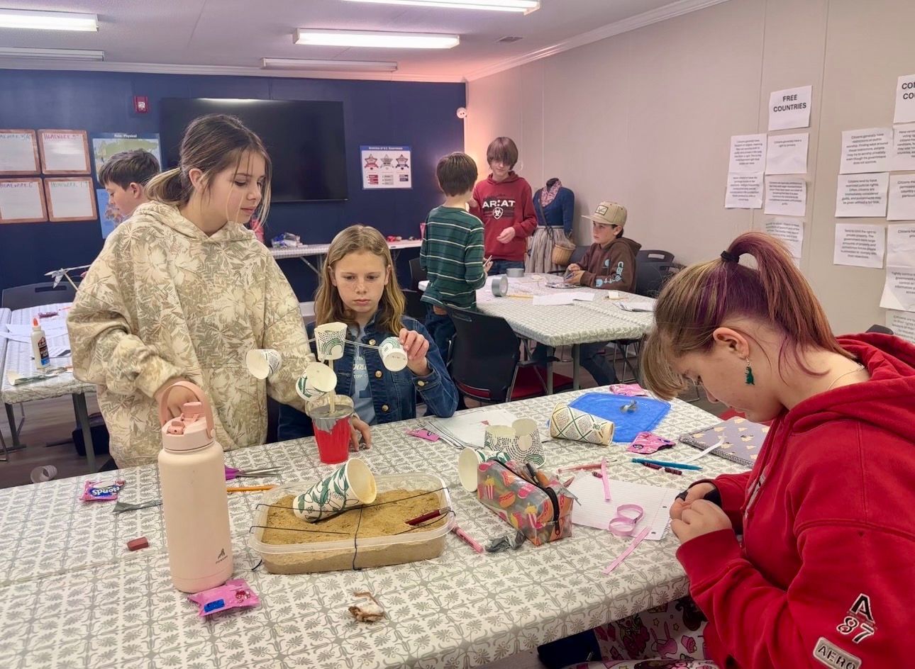 Children crafting at a table: papers, cups, and a water bottle visible. Indoor setting with others.