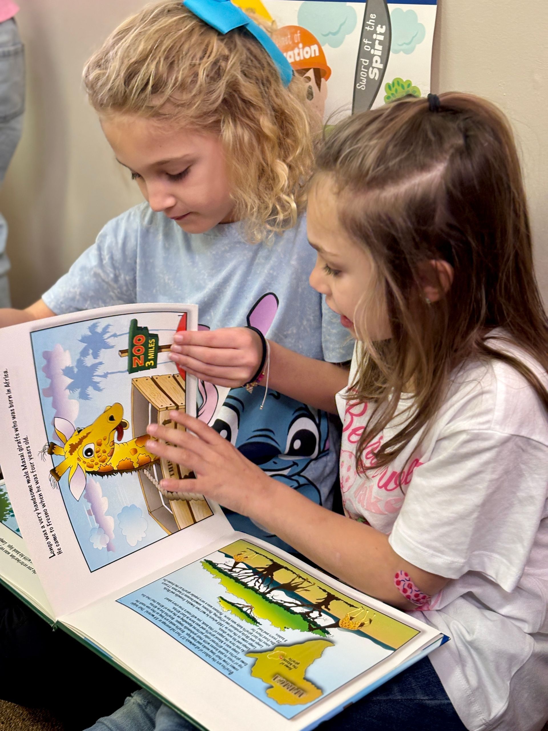 Two girls reading a book together; one points, colorful illustrations.