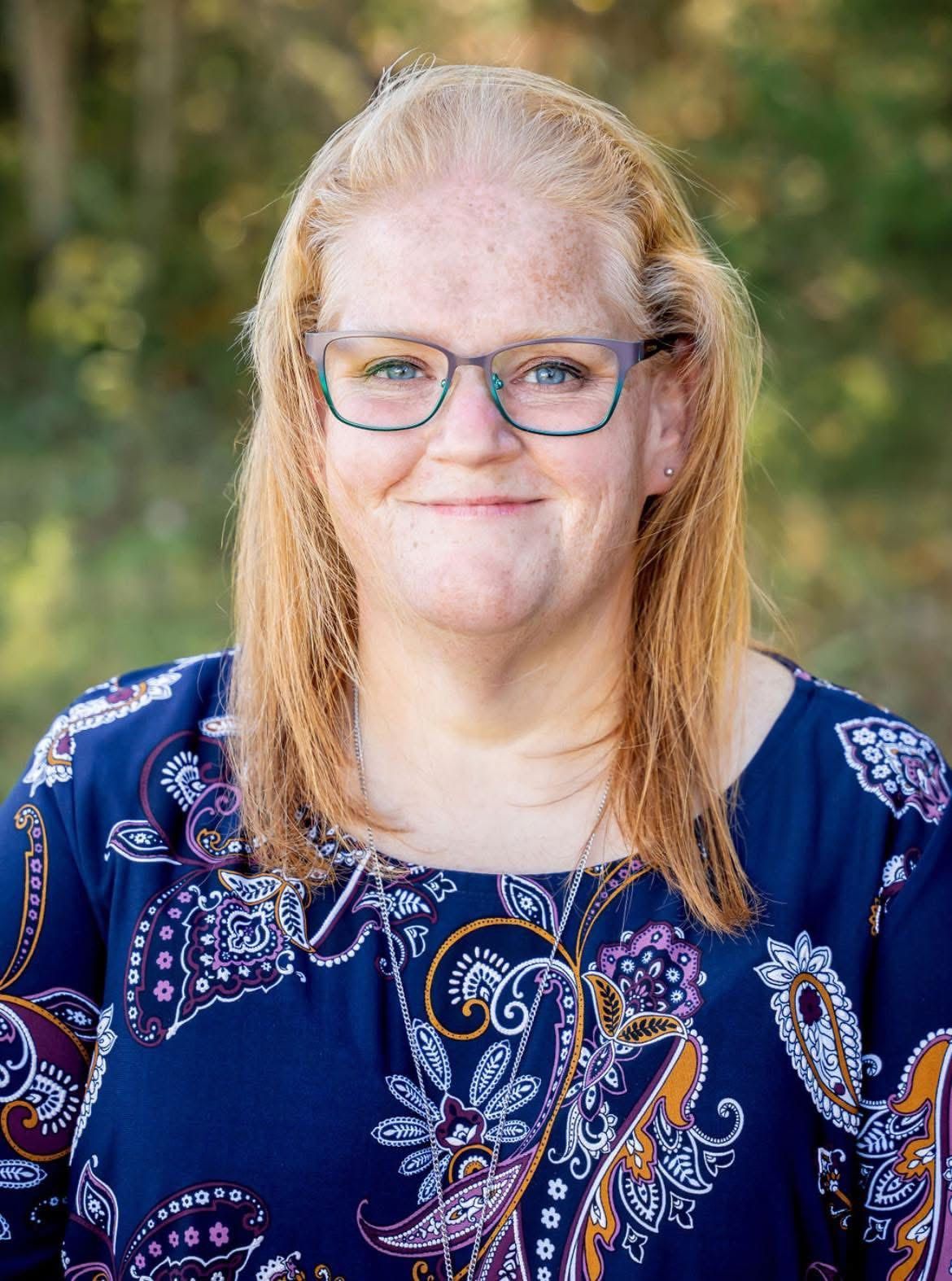 Woman with glasses, blond hair, and patterned blue top smiles outdoors.