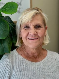 Woman with short blonde hair smiles, wearing a light gray ribbed sweater. Green plant on left.