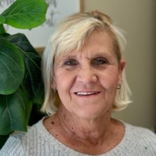 Woman with short blonde hair smiles, wearing a light gray ribbed sweater. Green plant on left.