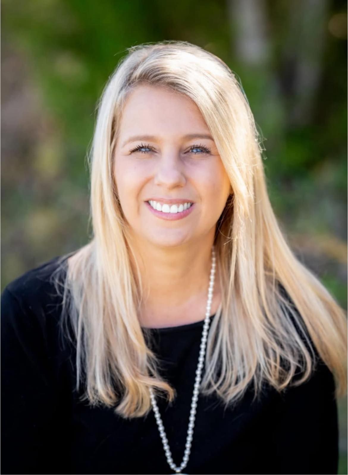 Blonde woman smiling, wearing a black top and pearl necklace, outdoors with greenery in the background.