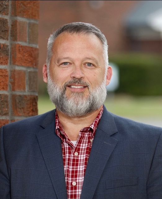 Man with graying beard, wearing a plaid shirt and blue blazer, smiling at camera near brick wall.
