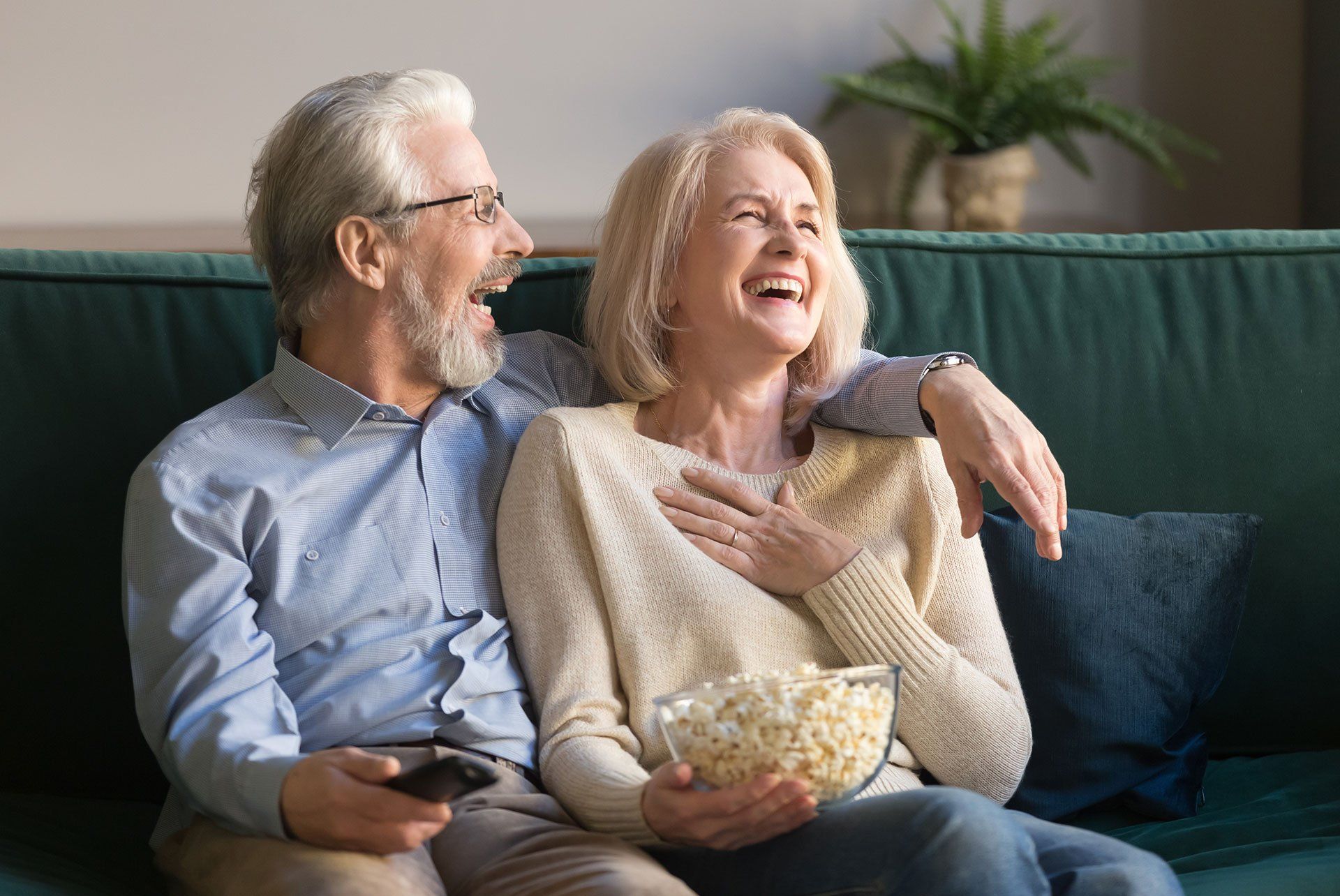 Laughing aged couple, man and woman watching tv, comedy show or movie and eating popcorn snack, sitting on cozy couch at home, mature family, man and woman enjoying free time, weekend together