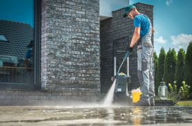 Worker pressure-washing a driveway beside a modern house, spraying water across the concrete. Worker pressure-washing a driveway beside a modern house, spraying water across the concrete.