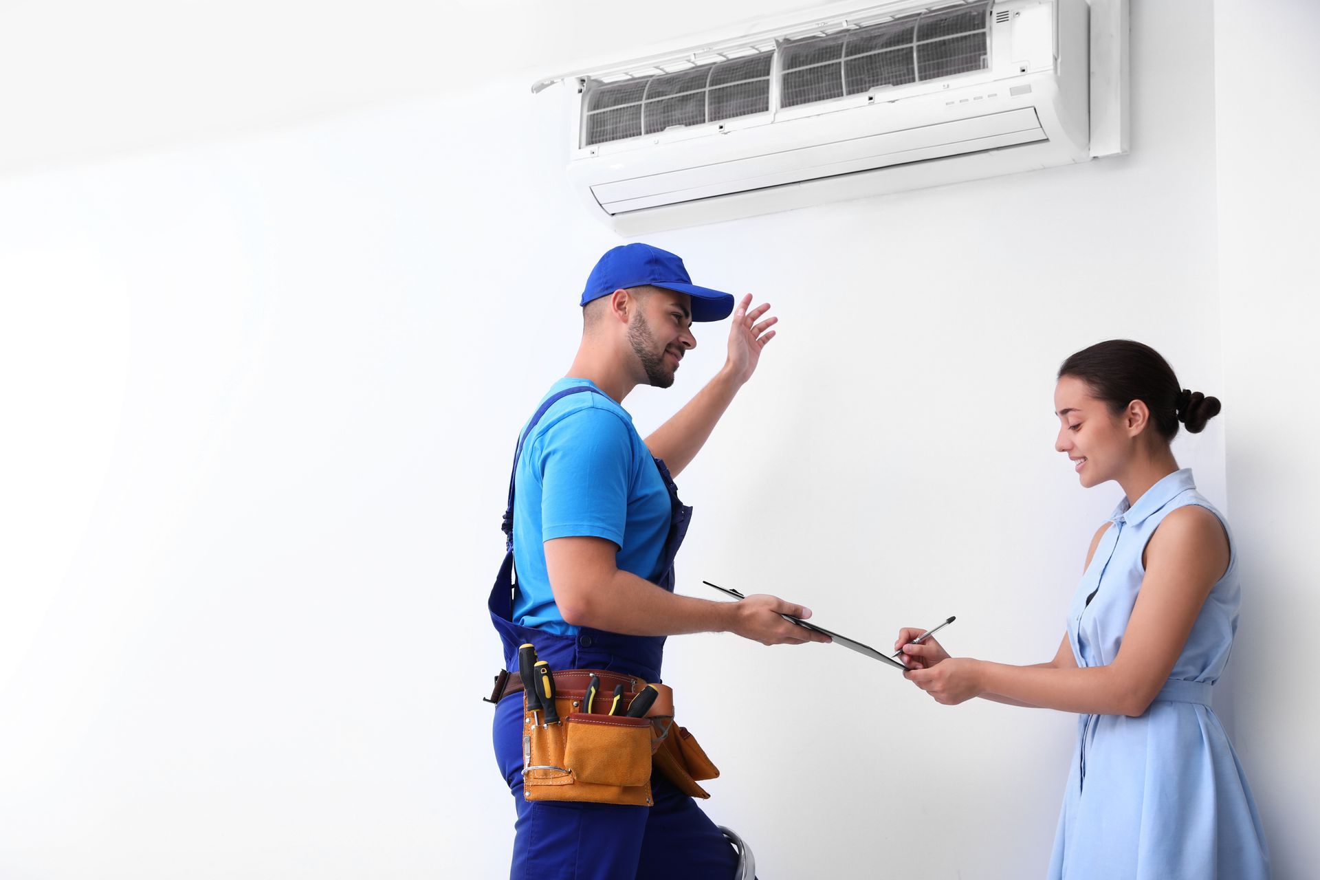 A technician wearing a blue uniform and tool belt points to a wall-mounted air conditioner while a client signs a form.