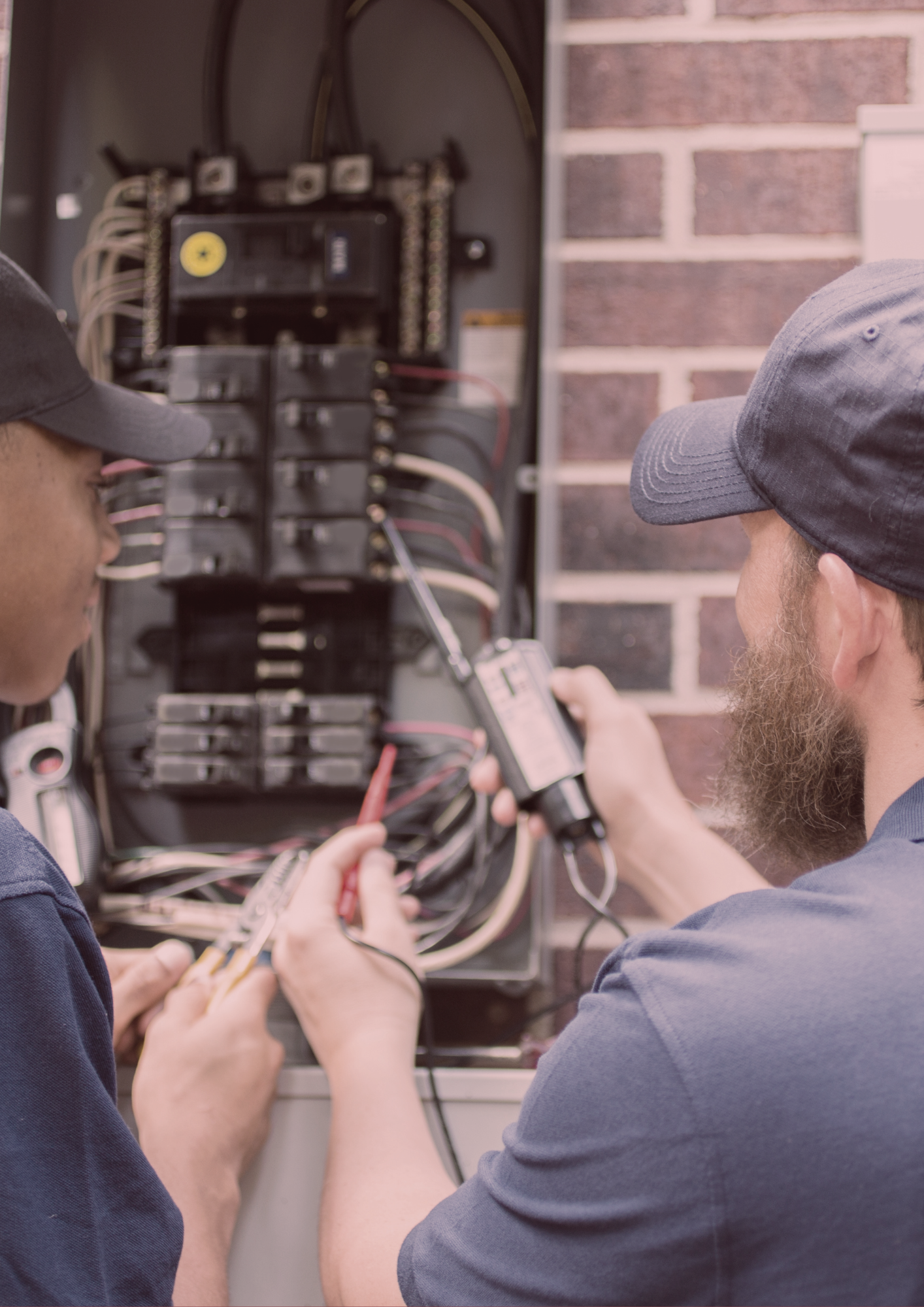 Two men are working on an electrical box with a brick wall in the background