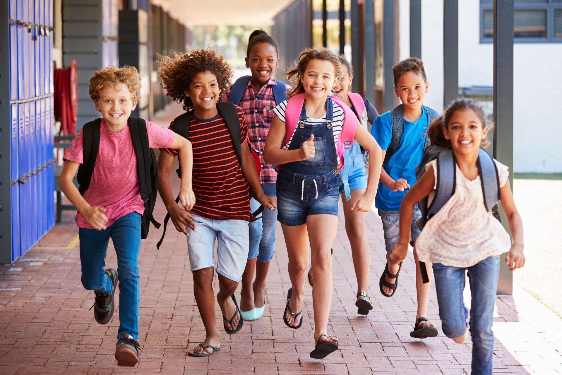 Group of children running down a school hallway, smiling and wearing backpacks.