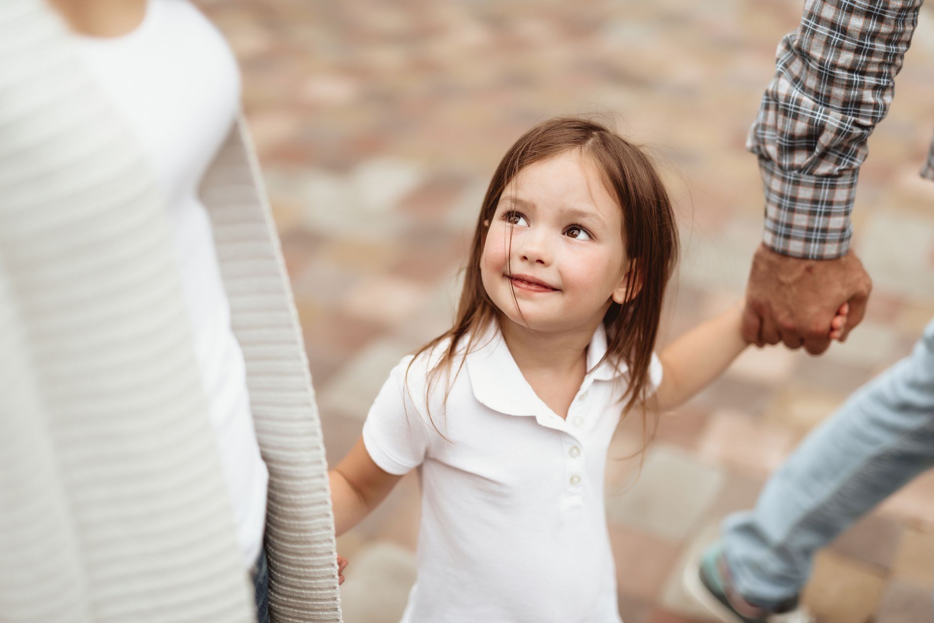 Child holding hands with two adults, looking up with a slight smile on a brick path.