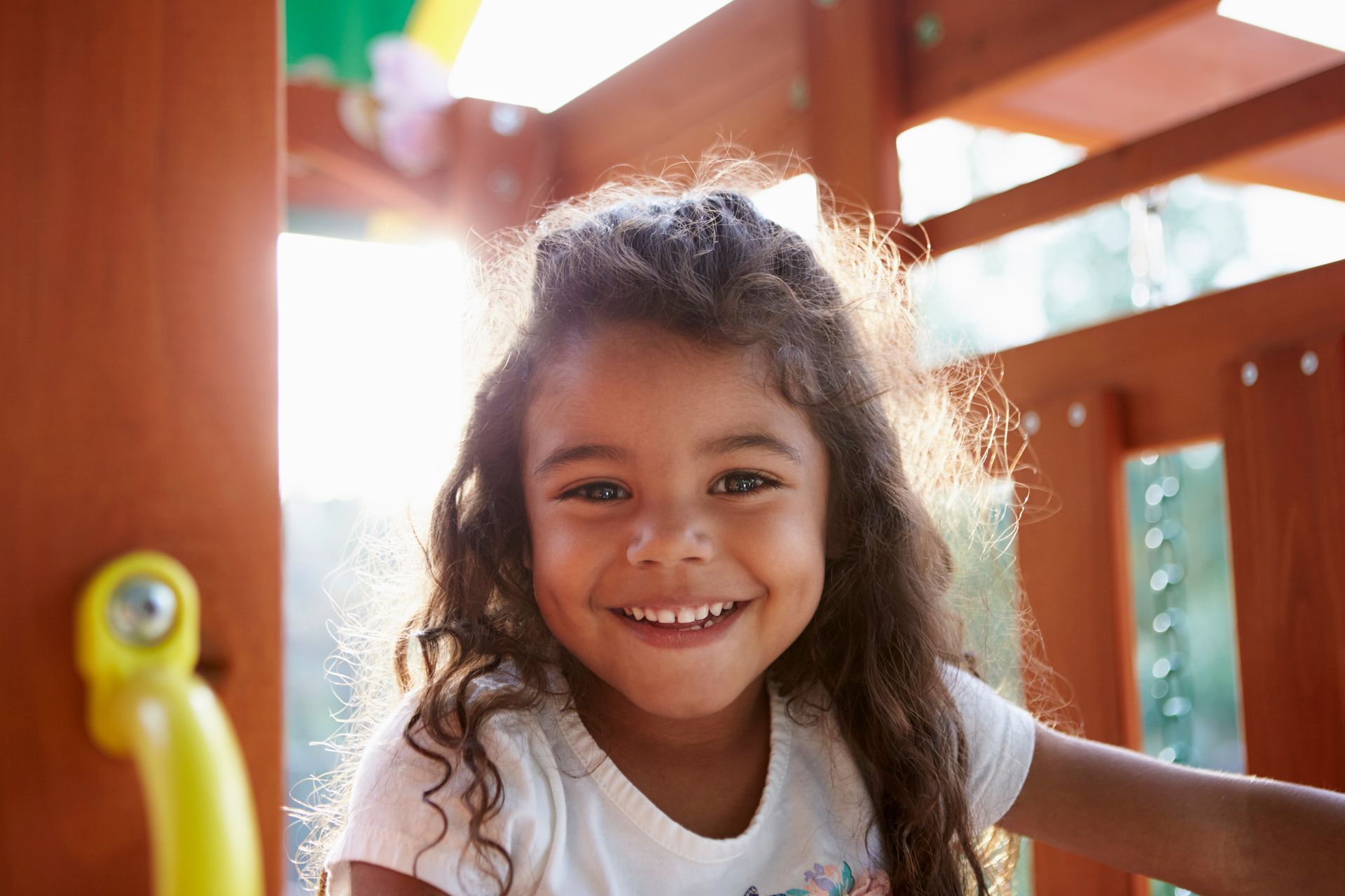 Smiling child on playground, long curly hair, bright sunlight.