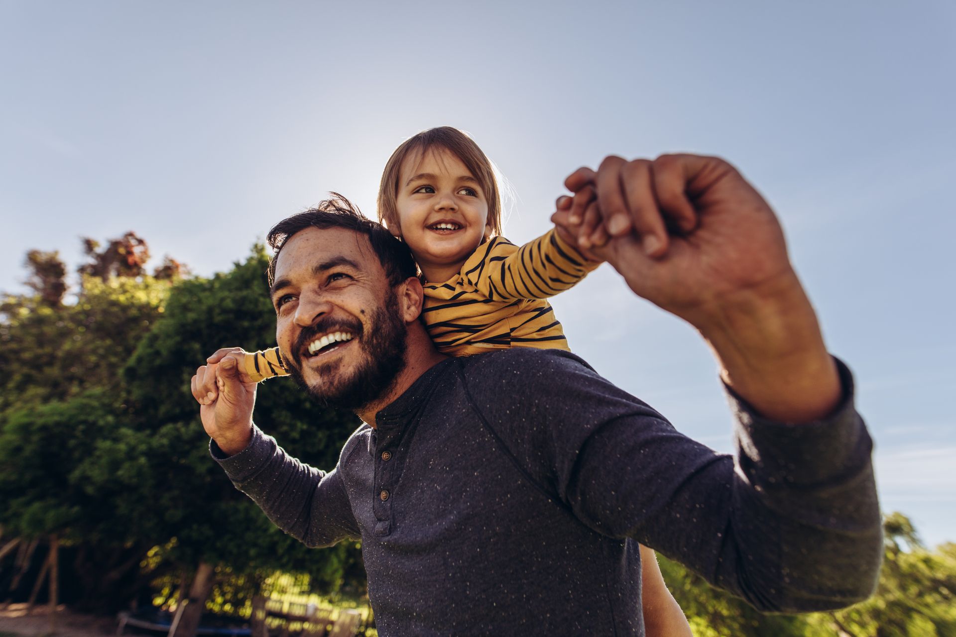 A man carries a smiling child on his shoulders outdoors, both looking up.