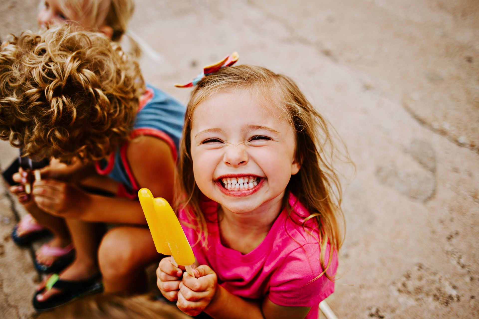 Girl with a pink shirt and bow smiles while holding a yellow popsicle. Two other children are nearby.