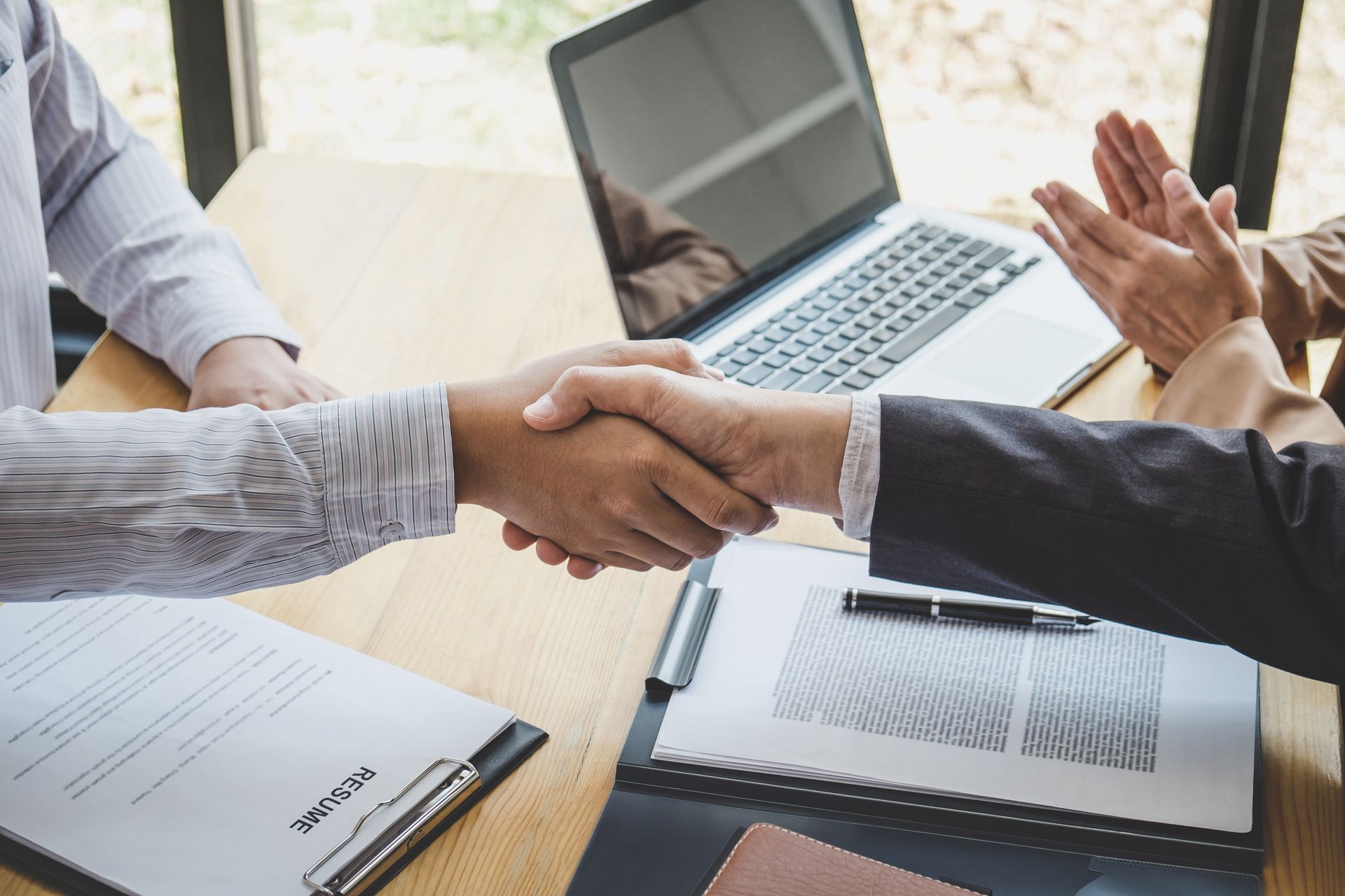Two people shaking hands, seated at a table with documents and a laptop, one person clapping.