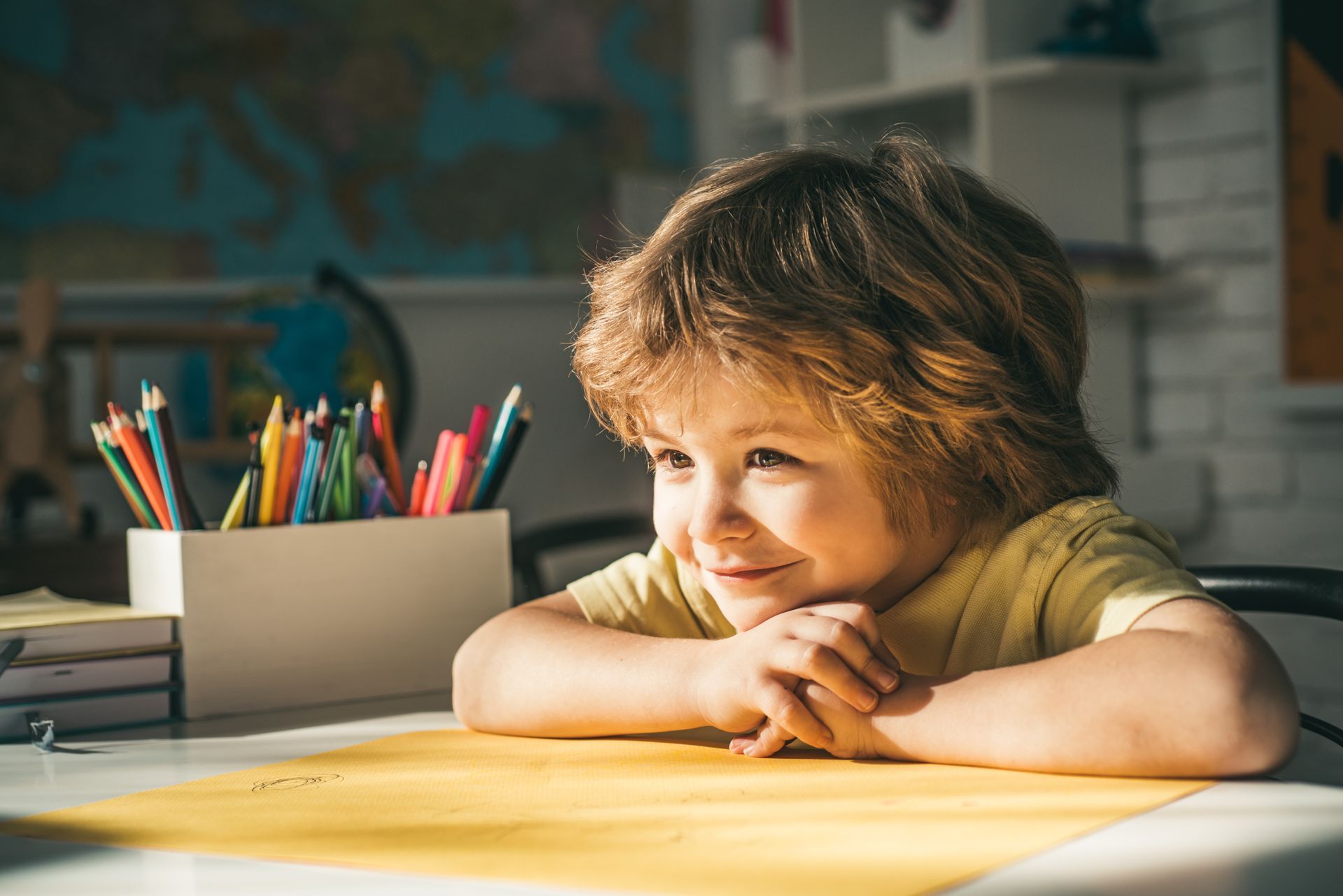 Boy with tousled hair, smiling, resting chin on hands at a desk with art supplies.